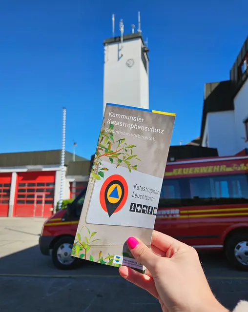 Hand hält Broschüre in Luft, im Hintergrund Feuerwehr und Feuerwehrauto zu sehen.