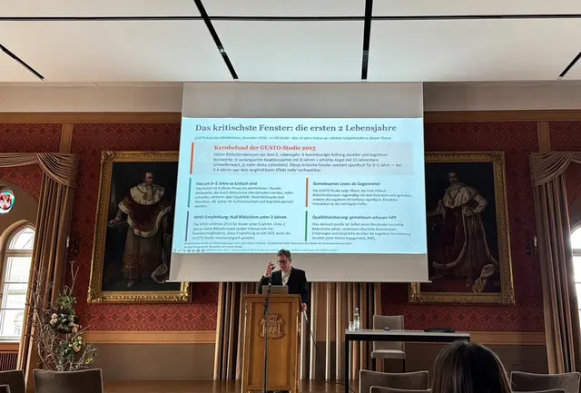 Referent am Pult im Festsaal, über ihm eine Leinwand mit Text