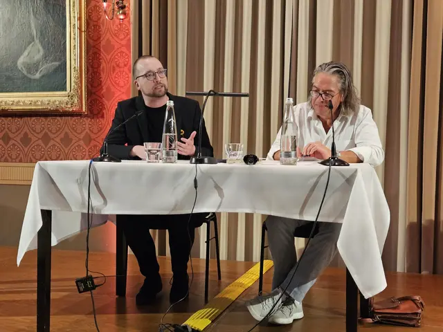 Zwei Männer auf einer Bühne im Festsaal an einem Tisch sitzend im Gespräch.
