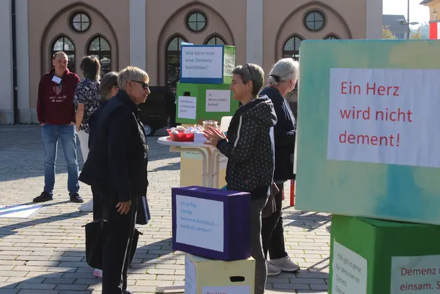 Personen unterhalten sich an einem Stand auf dem Wochenmarkt, im Vordergrund ein Spruch auf einem Zettel: Ein Herz wird nicht dement!
