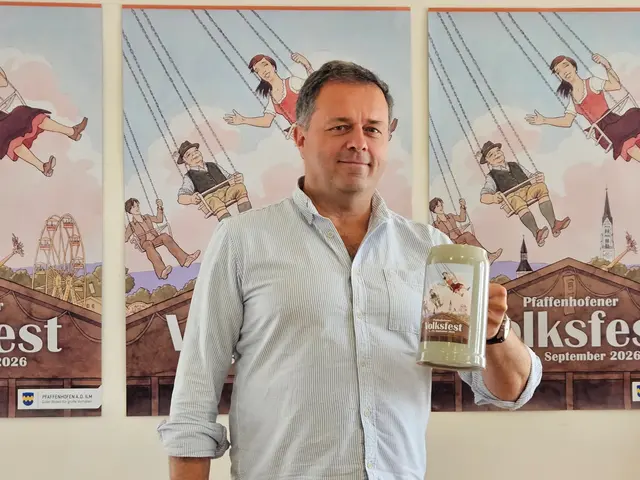 Gottfried Müller vor drei aufgehängten Plakaten, die er gestaltet hat und mit Maßkrug mit Volksfestmotiv in der Hand